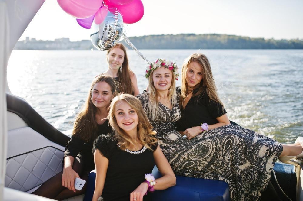 Five women celebrating bachelorette party on boat with balloons and floral crowns.