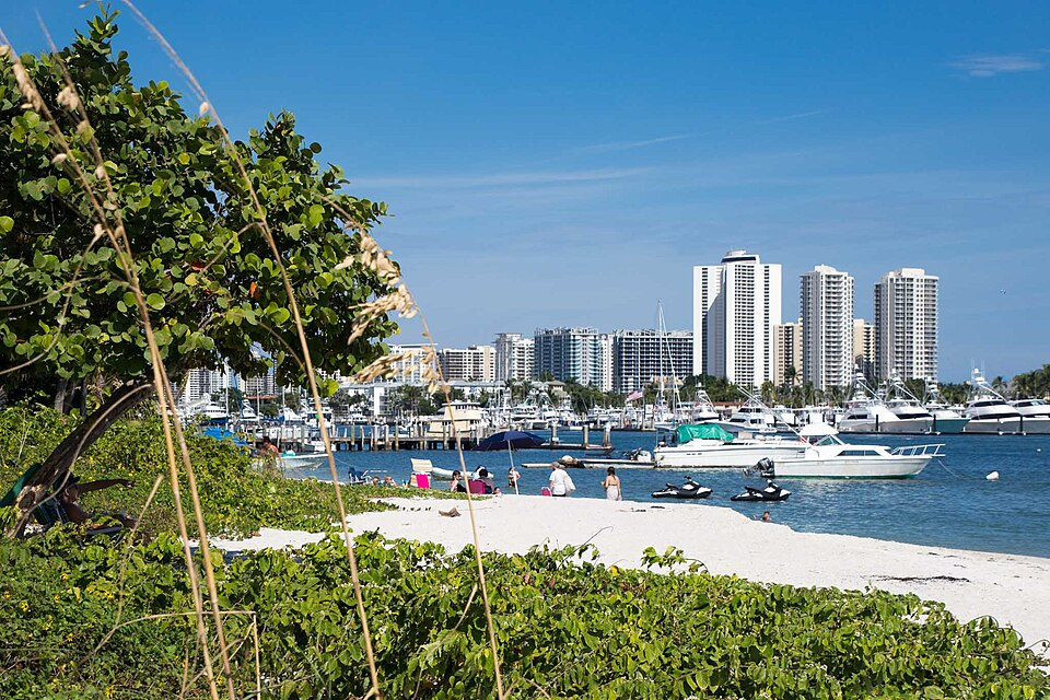 Peanut Island beach with boats, skyline, and tropical greenery.